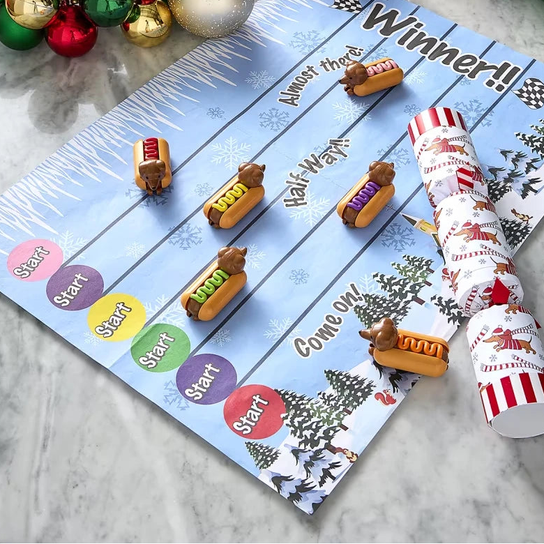 A festive Christmas-themed hot dog race game laid out on a marble surface, featuring a blue scoring board with colorful start/finish zones, several wooden hot dogs on tracks, two rolled-up red-and-white party crackers, and holiday ornaments around the edges.