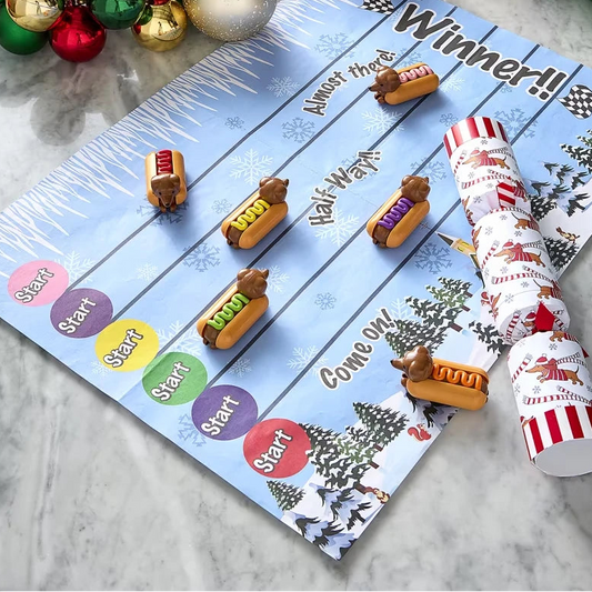 A festive Christmas-themed hot dog race game laid out on a marble surface, featuring a blue scoring board with colorful start/finish zones, several wooden hot dogs on tracks, two rolled-up red-and-white party crackers, and holiday ornaments around the edges.