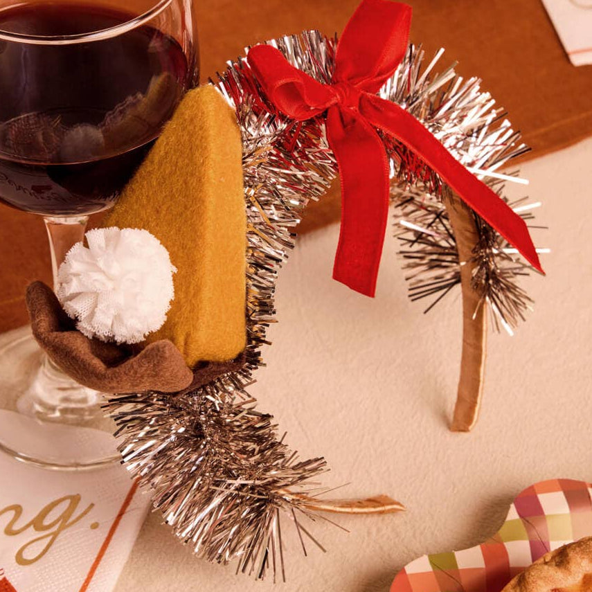 A festive holiday tabletop scene featuring a silver tinsel garland with a red ribbon bow, a small brown napkin with a white pom-pom, and a yellow pastry, alongside a glass of red wine and pie in the background.