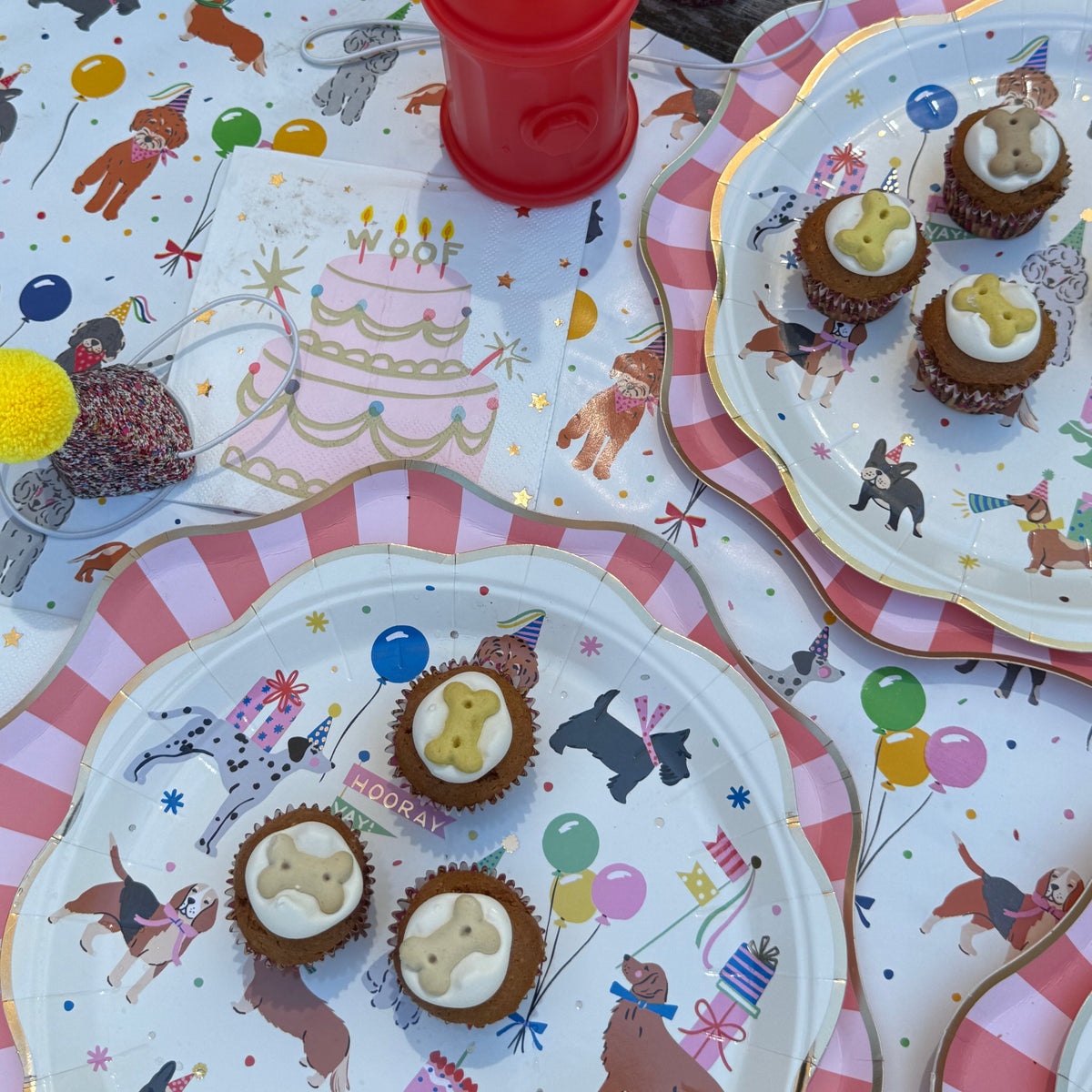 A styled birthday party table setting featuring printed paper plates and a matching tablecloth with a dog-themed circus design, topped with cupcakes and a red party hat.