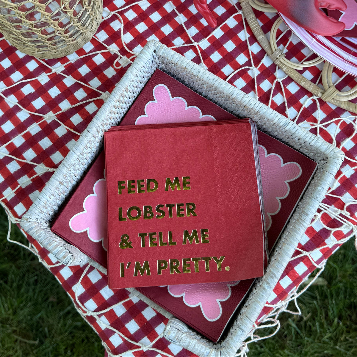 A red lobster figurine and a red square sign with gold text reading 'FEED ME LOBSTER & TELL ME I'M PRETTY.' are arranged on a red-and-white checkered picnic blanket outdoors on grass, with woven baskets, plates, and tomatoes visible around them.