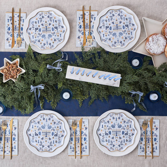 Hannukah table setting with decorative plates, cutlery, and greenery on a blue runner.