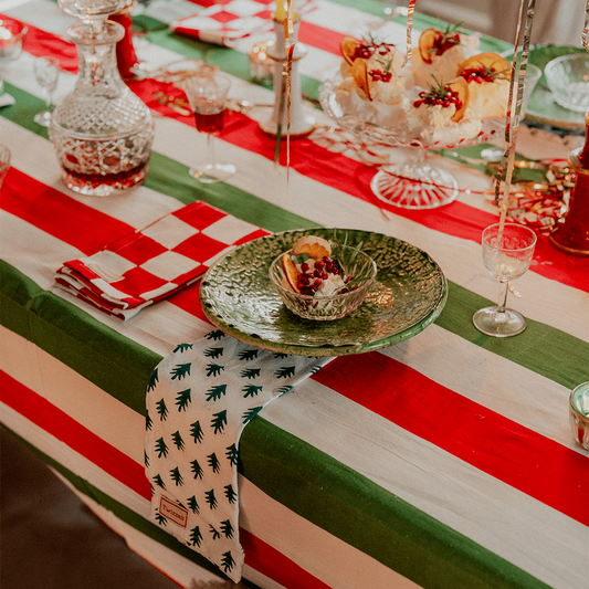 A festive holiday table setting featuring a tablecloth with bold red, green, and white stripes, accented with a patterned runner, checkered napkins, and glassware.