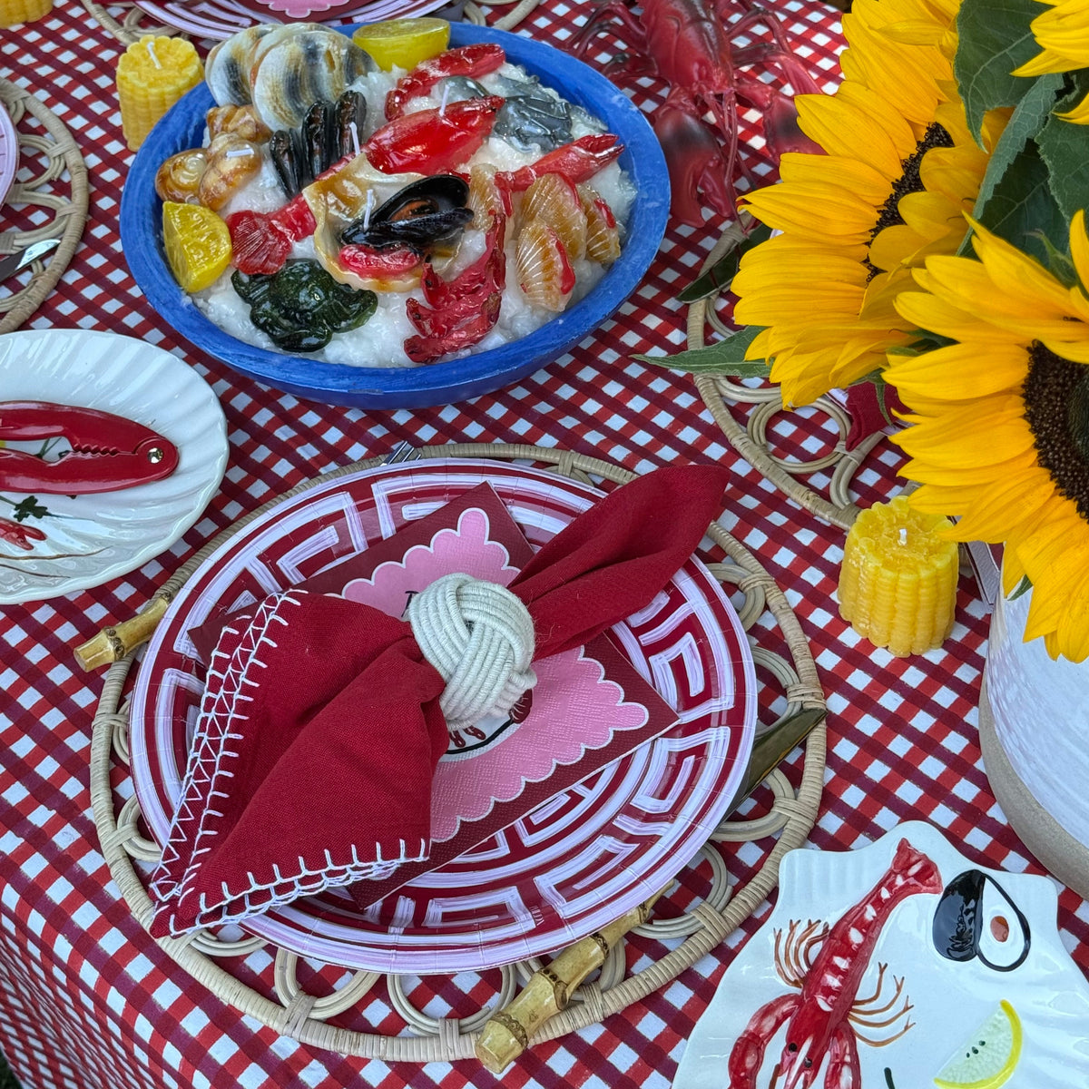 Outdoor dining table setting with a red and white gingham tablecloth, matching plates, red napkins with white stitching, a blue bowl of seafood, and large yellow sunflowers.