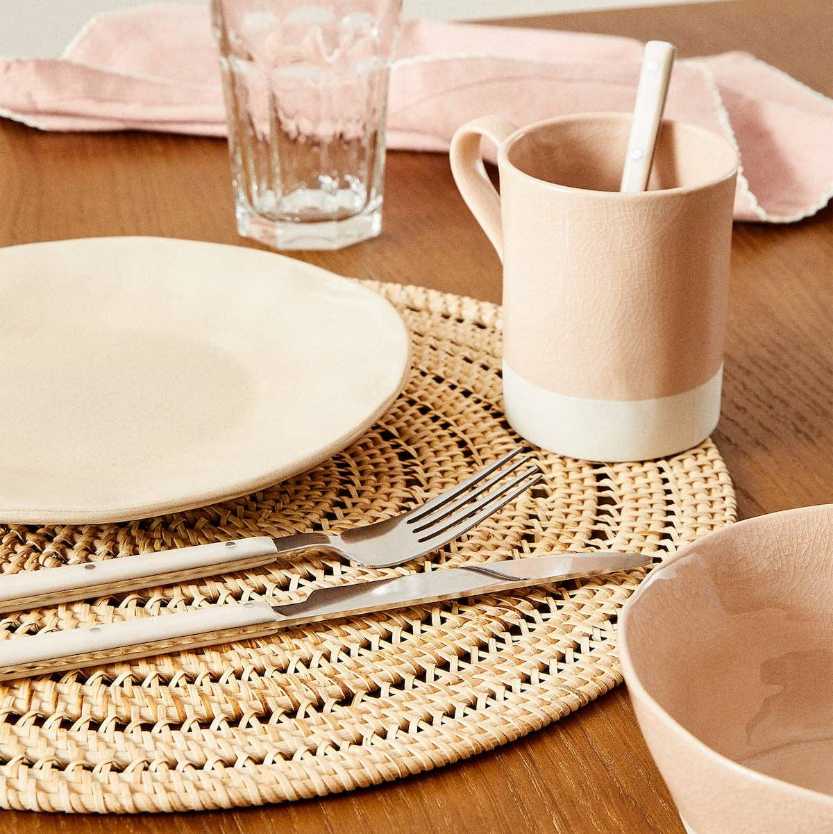 A styled dining table setting featuring a light pink ceramic mug with a white base and a spoon inside, alongside a white plate, a woven placemat, silverware, a glass, and a pink napkin on a wooden table.