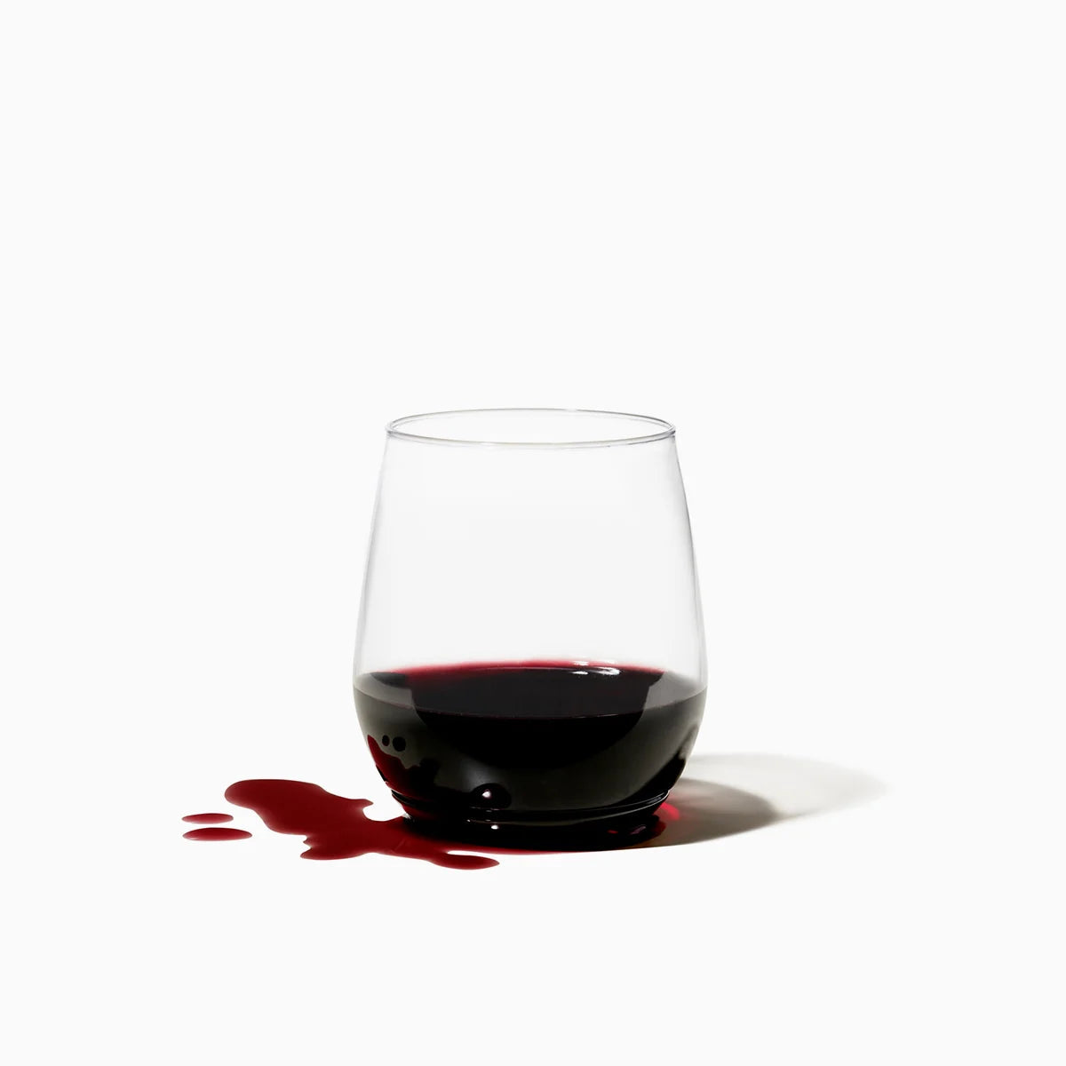 A clear stemless wine glass containing a small amount of dark red wine, with a red liquid spill on a white surface against a white background.