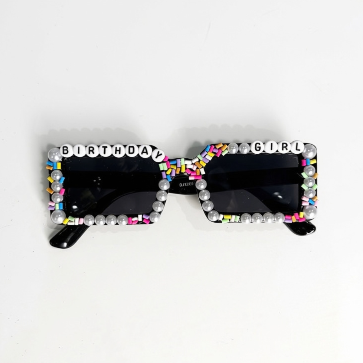 Black square sunglasses with a decorative border of white faux pearls and colorful letter tiles spelling 'BIRTHDAY GIRL' across the top and bottom, shown on a plain white background.