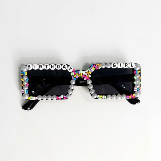 Black square sunglasses with a decorative border of white faux pearls and colorful letter tiles spelling 'BIRTHDAY GIRL' across the top and bottom, shown on a plain white background.