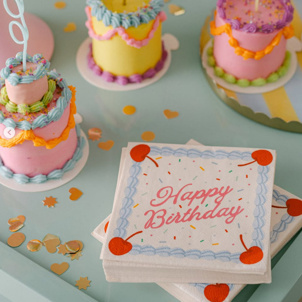 A birthday party scene with three small decorated  mini cakes on a pastel mint table, scattered gold confetti, and a stack of white napkins in the foreground printed with 'Happy Birthday' and cherries.