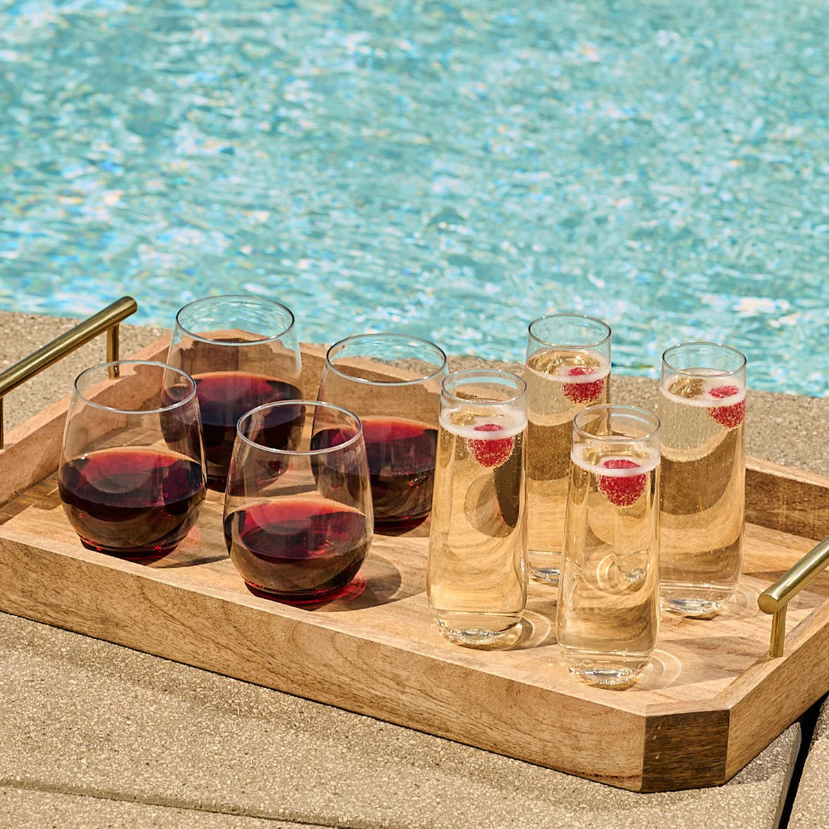 A wooden serving tray with several clear drinking glasses, including two filled with red wine and four with a pale golden beverage topped with raspberries, set beside a swimming pool.