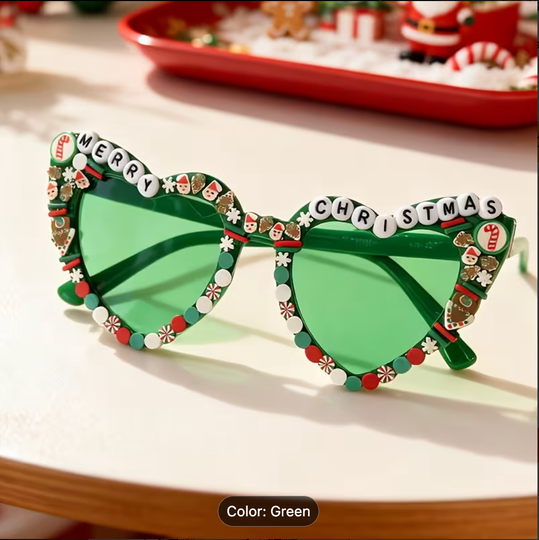 Green heart shaped sunglasses with decorative holiday embellishments, including bead borders, candy cane details, and white letter beads spelling 'MERRY CHRISTMAS' on the upper frame, shown on a tabletop with a blurred festive background.