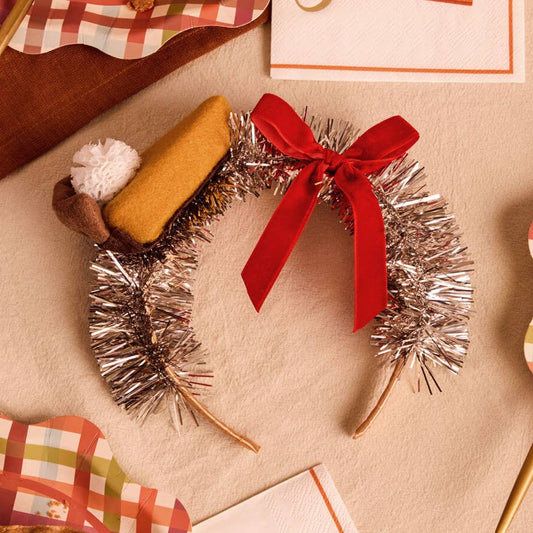 A festive tinsel headband styled with a red ribbon, a mini pumkin pie topper, and a small white pom-pom, arranged on a beige surface with autumn-themed paper napkins and pie slices.