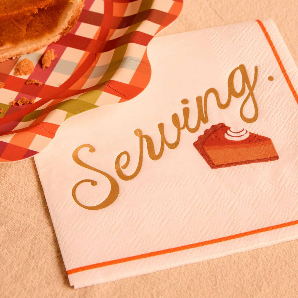 A close-up of a white paper napkin printed with the word "Serving." in gold script and a small pie slice illustration, styled with a partially visible pumpkin pie on a checkered napkin liner over a beige surface.