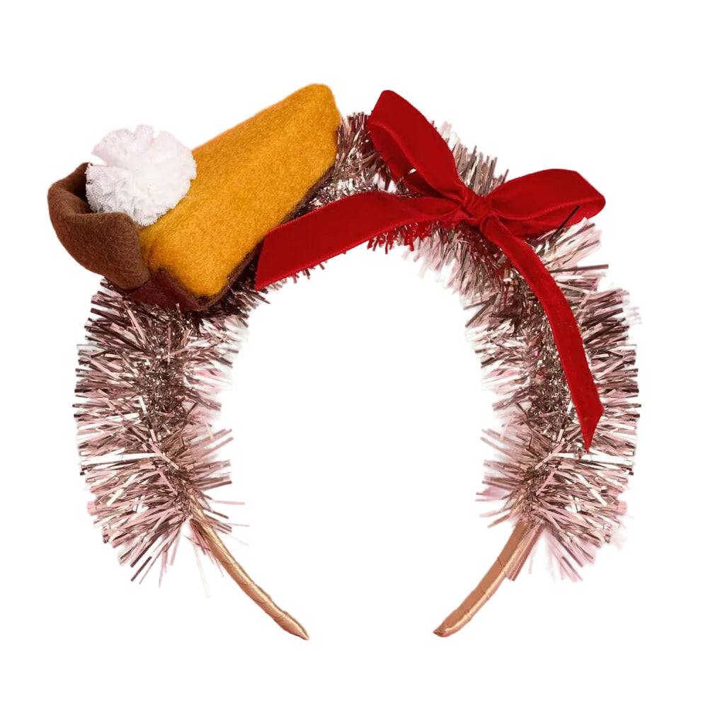 A novelty headband styled like a slice of pumpkin pie, featuring a metallic silver tinsel arch with a large red ribbon bow, a mustard-yellow felt slice, a brown crust edge, and a white whipped cream pom-pom on a pink background.