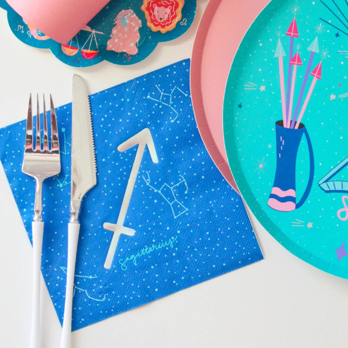 A styled flat-lay of astrology-themed tableware featuring a blue napkin with a white sword constellation and the word 'Sagittarius', a pink plate, a turquoise plate with a bow and arrow illustration and the word 'Sagittarius', a pink rolled napkin, and silver cutlery on a white surface.
