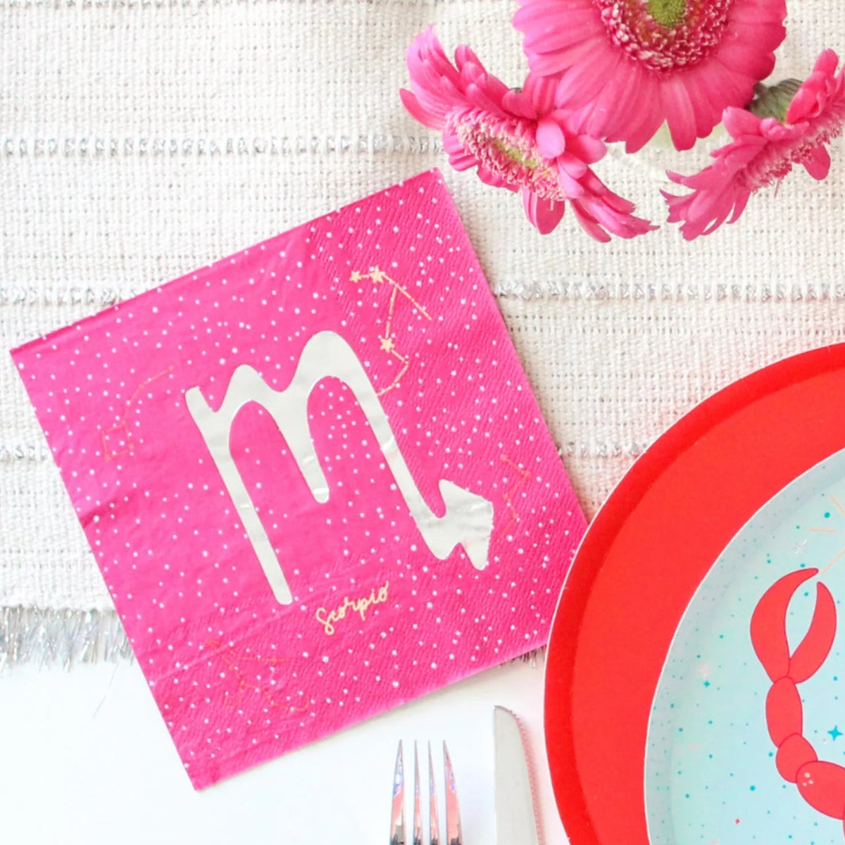 A styled flat-lay of pink paper napkins featuring the Scorpio zodiac symbol and stars, alongside a red-rimmed plate with a crab illustration, a fork, a knife, and pink flowers on a textured white fabric background.