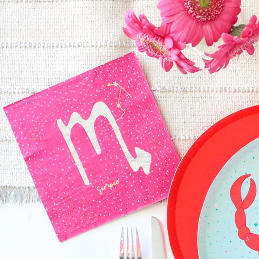 A styled flat-lay of pink paper napkins featuring the Scorpio zodiac symbol and stars, alongside a red-rimmed plate with a crab illustration, a fork, a knife, and pink flowers on a textured white fabric background.