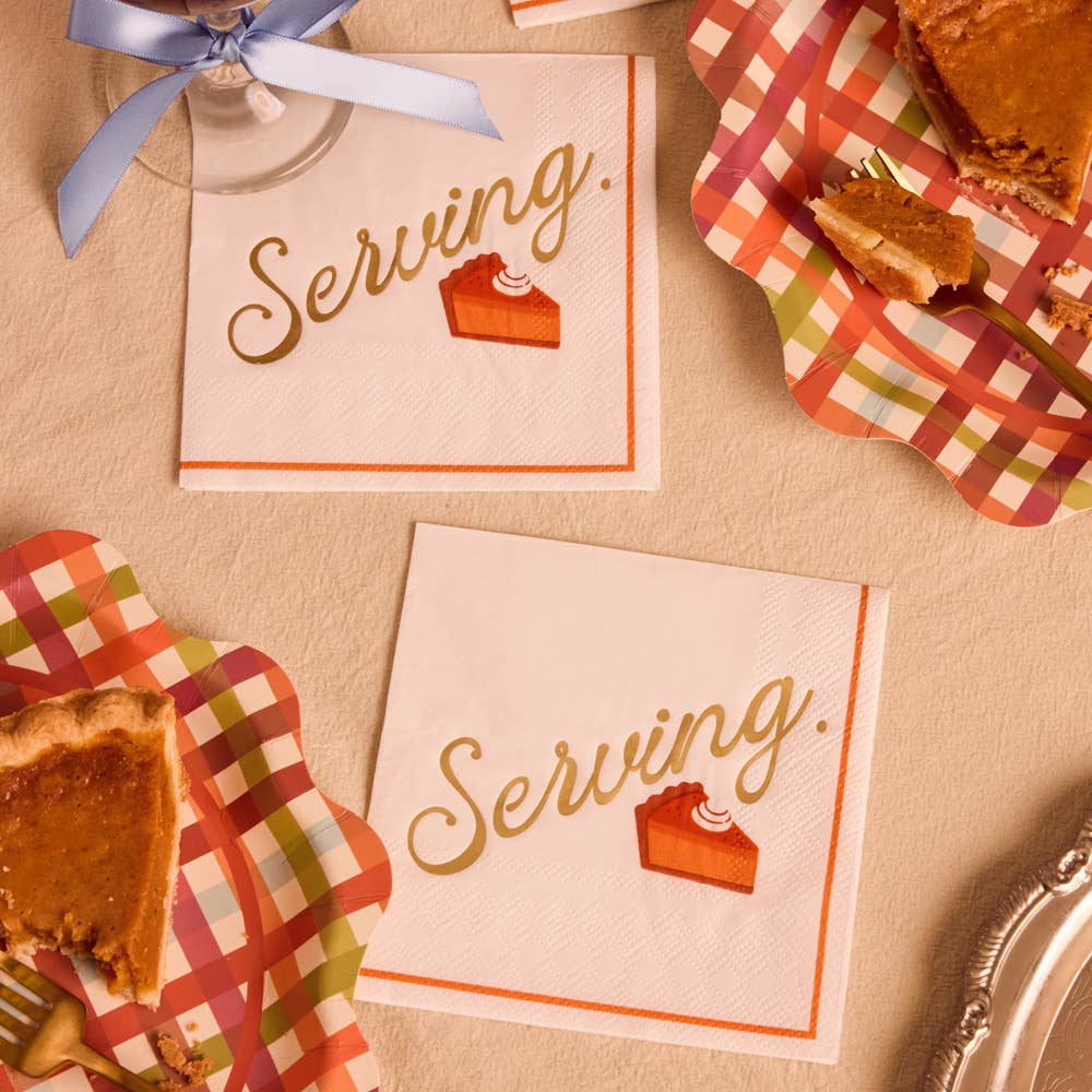 Two white paper napkins with 'Serving.' in gold script and a small pumpkin pie graphic, styled on a beige tablecloth with pie slices and plaid placemats.