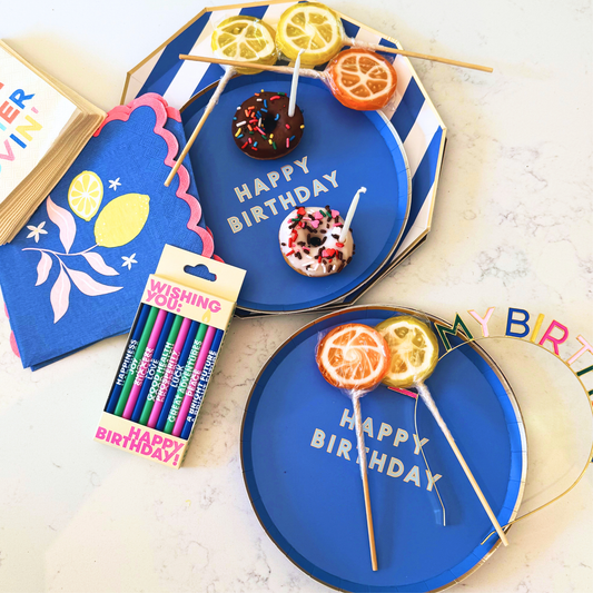 Birthday party supplies set including blue 'HAPPY BIRTHDAY' paper plates, matching blue napkins, colorful 'Wishing Fab' markers, and decorative orange and lemon slices, arranged on a light marble surface.