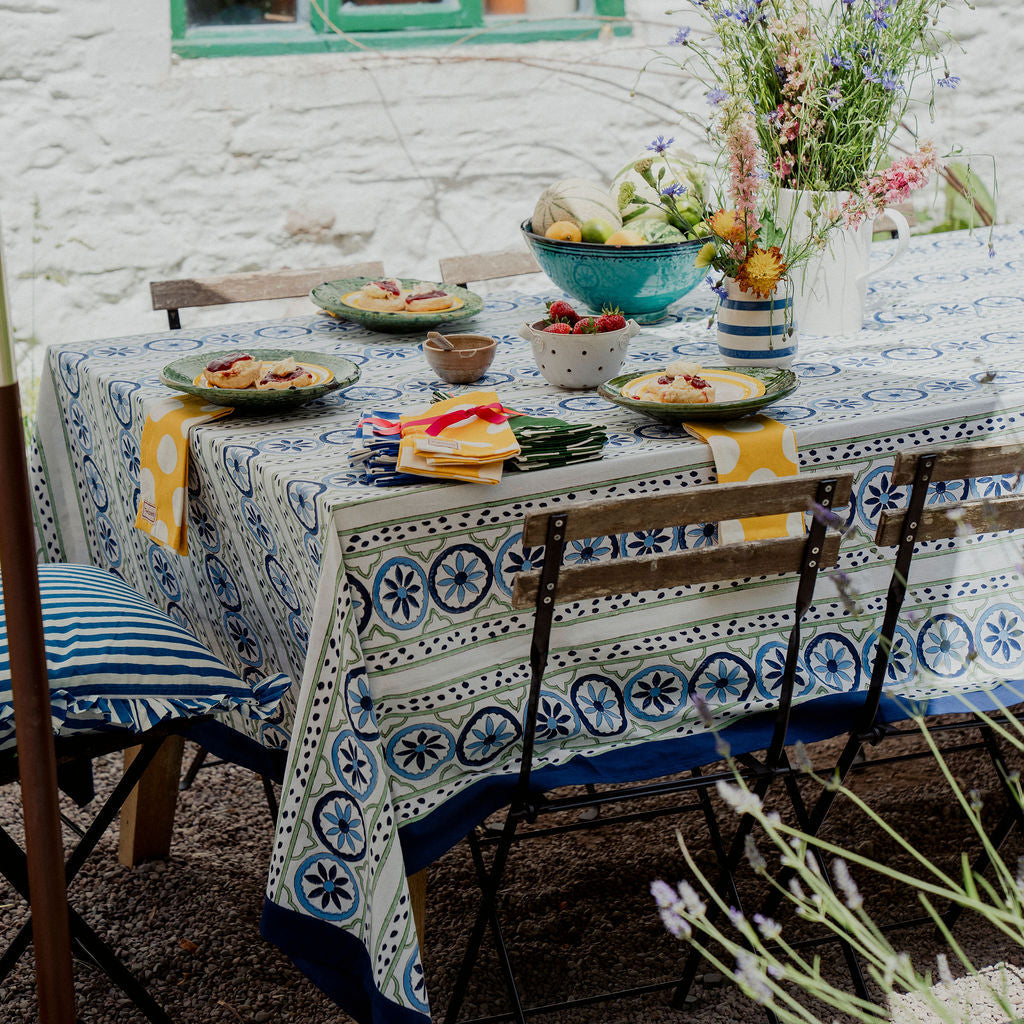 A rustic outdoor dining table set with a blue and white patterned tablecloth, matching plates, yellow napkins, and a large floral centerpiece against a white brick wall with a green window.