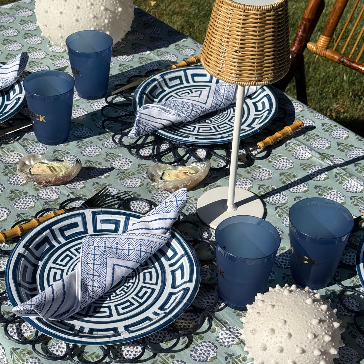 Outdoor dining table set with blue and white patterned plates, matching blue napkins, blue cups, a wicker lamp, and white textured sea urchin light decor on a patterned tablecloth.