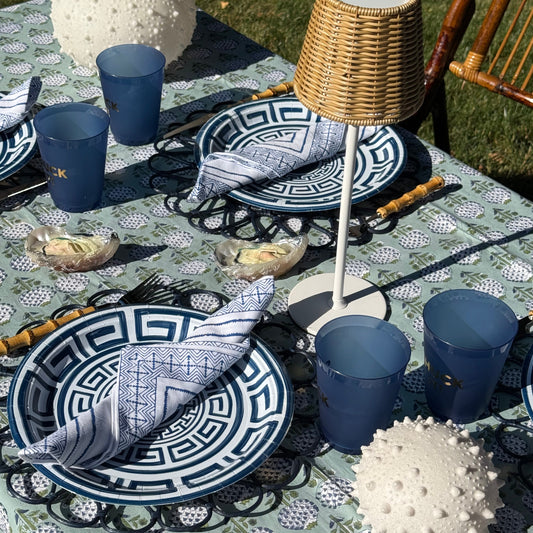 Outdoor dining table set with blue and white patterned plates, matching blue napkins, blue cups, a wicker lamp, and white textured sea urchin light decor on a patterned tablecloth.