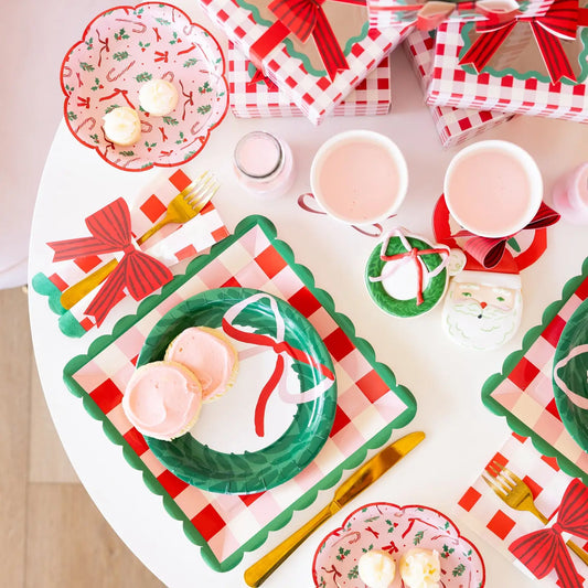 A festive holiday table setting featuring red and green gingham placemats, matching paper plates, napkins, and gift boxes with red bows, styled with pink frosted cupcakes and white mugs of iced coffee on a white round table.