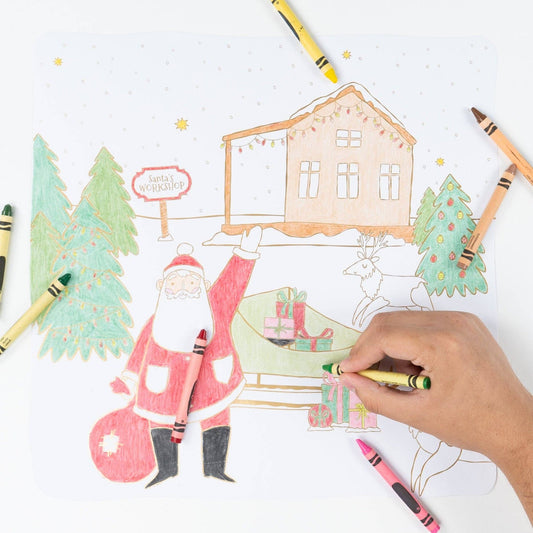 A hand is coloring a Christmas-themed drawing with crayons on a white surface. The drawing shows Santa Claus, a house, trees, gifts, and a reindeer, with several crayons placed around it.