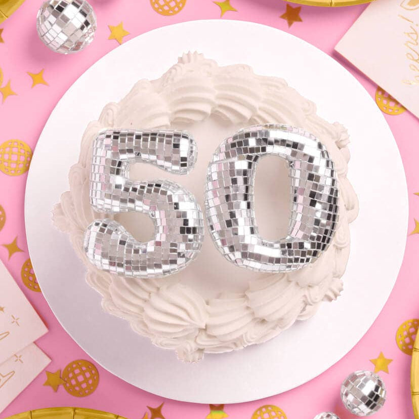 White cake with silver disco ball numbers '50' on a pink background with decorations.