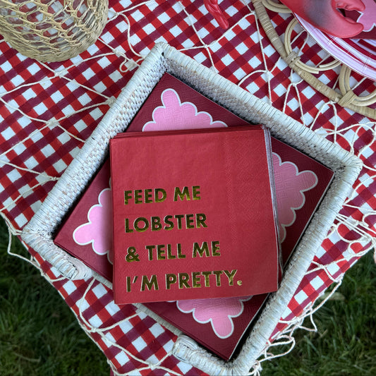 A red lobster figurine and a red square sign with gold text reading 'FEED ME LOBSTER & TELL ME I'M PRETTY.' are arranged on a red-and-white checkered picnic blanket outdoors on grass, with woven baskets, plates, and tomatoes visible around them.