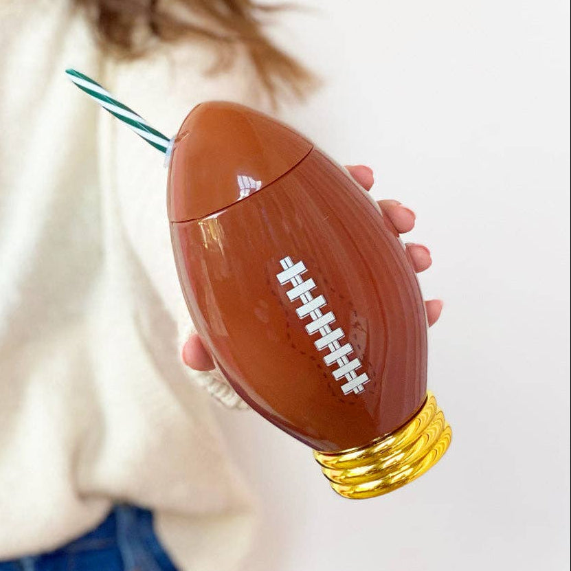 Person holding a football-shaped tumbler with a straw on a white background