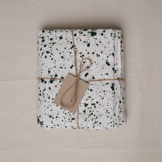 A folded white tablecloth with a green speckled pattern, wrapped in twine with a kraft paper tag, placed on a neutral beige fabric background.