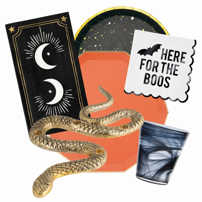 Halloween party tableware set featuring a gold snake, a black and gold tarot card plate, an orange paper plate, a white napkin with a bat and 'HERE FOR THE BOOS' print, and a black and white marbled cup on a white background.