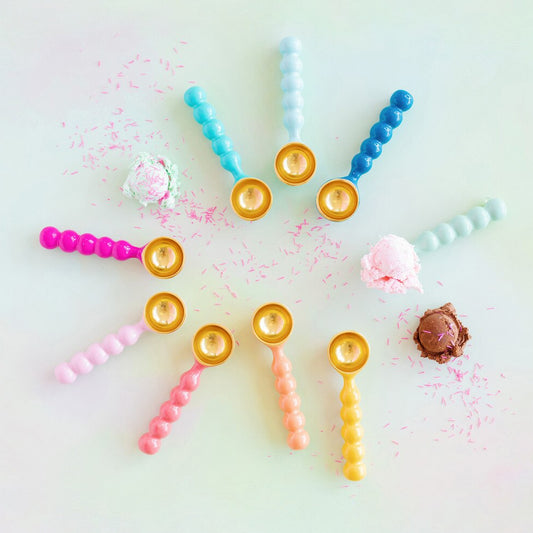 Nine enameled ice cream scoops arranged in a circular pattern on a pale mint background, with small decorative sprinkles and ice cream scoops around them. The scoops come in pastel pink, peach, yellow, blue, and aqua, each with a gold-toned circular end.