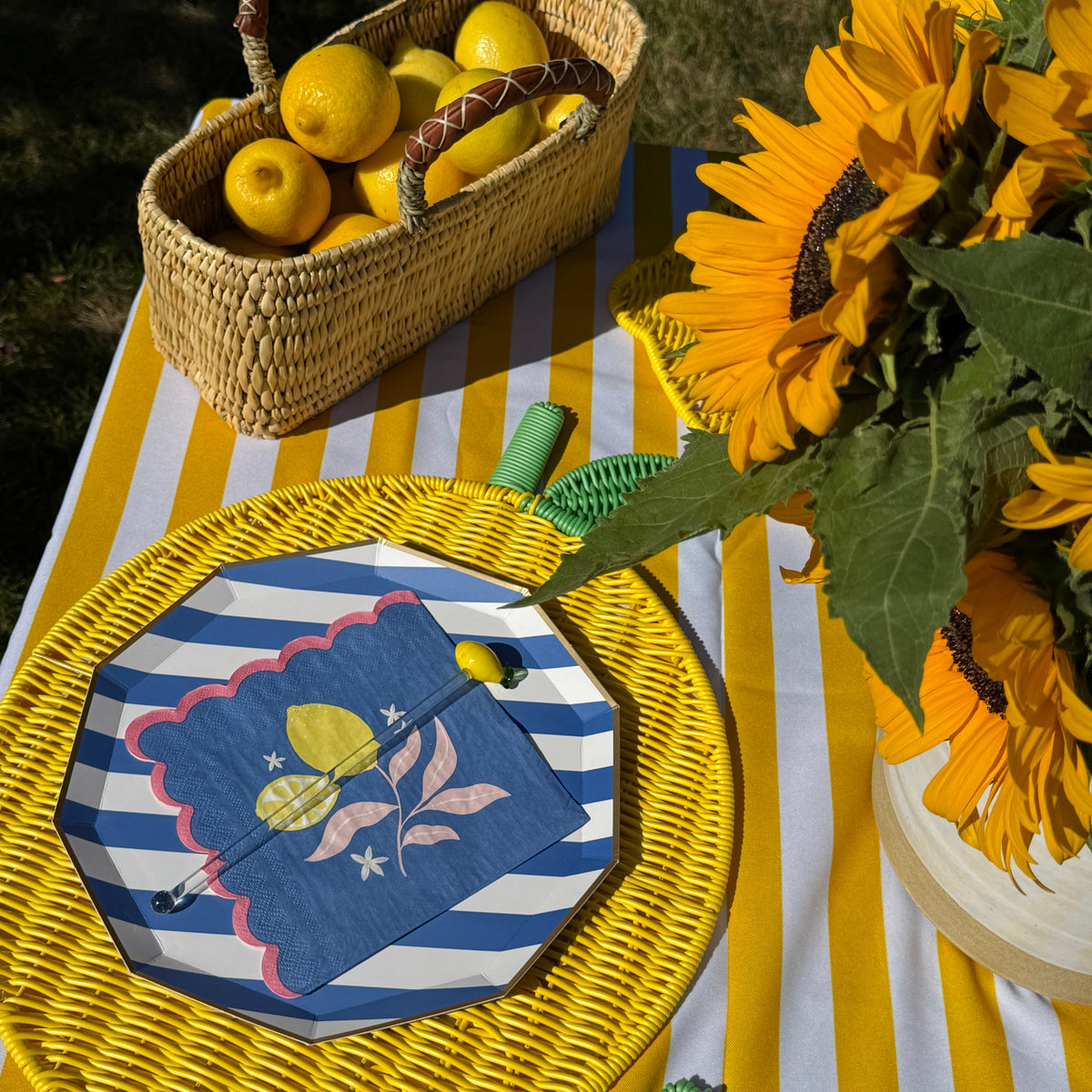 Outdoor picnic-style table setting with a woven basket of lemons, a yellow sunflower bouquet, and a yellow-and-white striped tablecloth. A round woven placemat holds a blue-and-white striped napkin with a yellow lemon illustration and a small spoon.