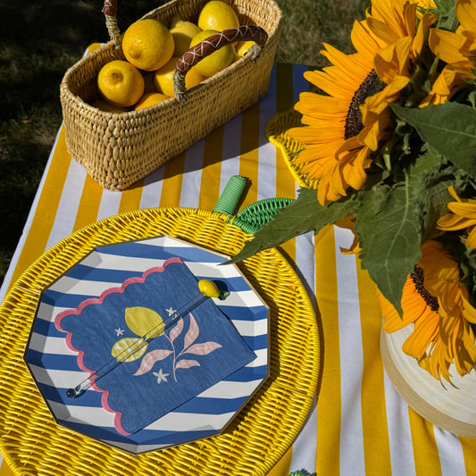Outdoor picnic-style table setting with a woven basket of lemons, a yellow sunflower bouquet, and a yellow-and-white striped tablecloth. A round woven placemat holds a blue-and-white striped napkin with a yellow lemon illustration and a small spoon.