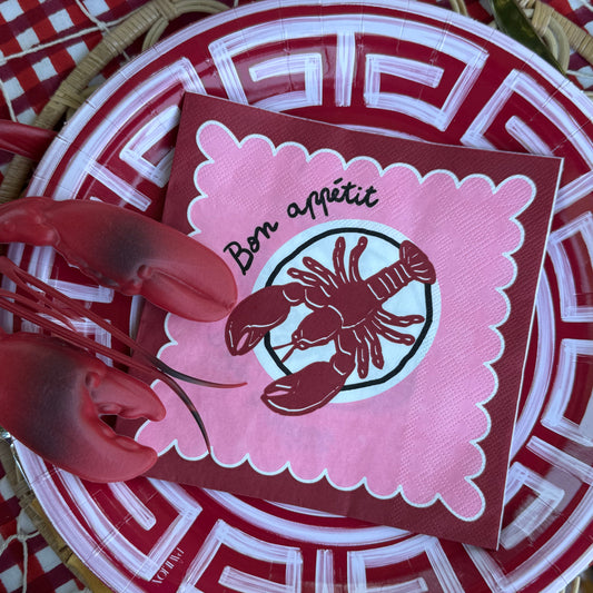 A close-up of a red and pink lobster-themed place setting, featuring a pink napkin with 'Bon appétit' and a lobster illustration, layered on a red and white patterned paper plate over a woven placemat.