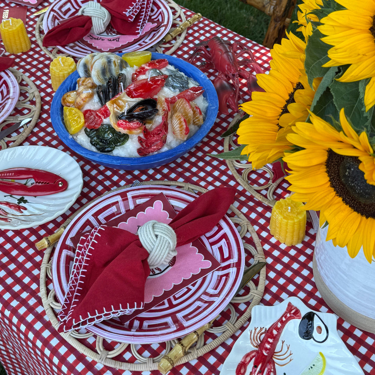 Outdoor dining table setup with a red and white gingham tablecloth, red greek key patterned plates, red lobster napkins, a blue bowl of seafood salad, yellow candles, and a large vase of sunflowers.