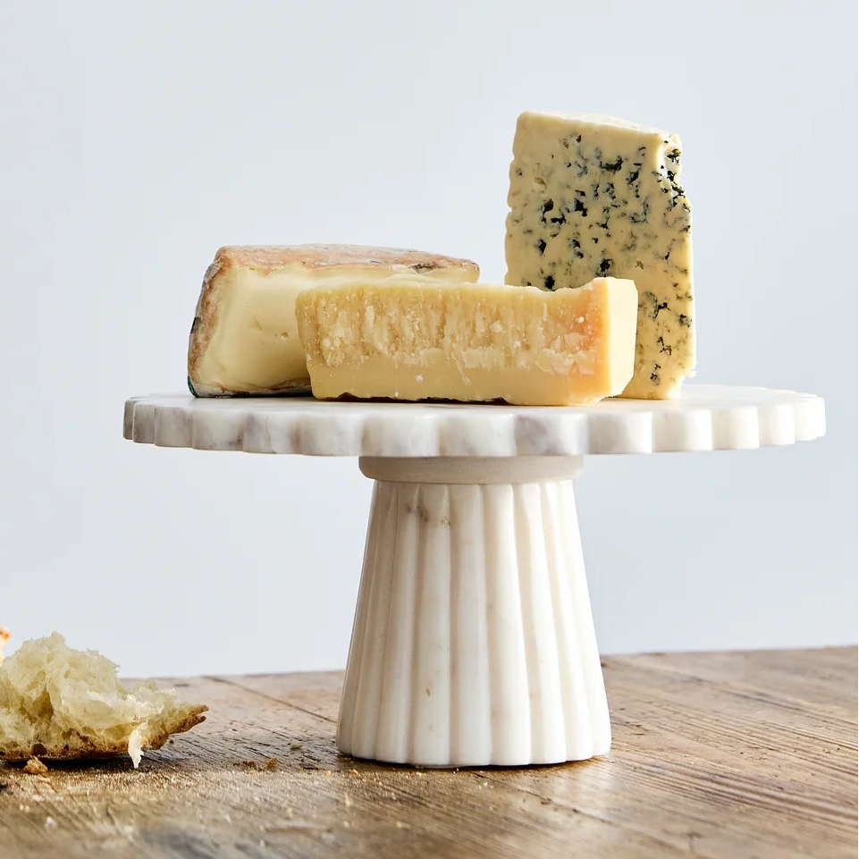 A white ceramic fluted cake stand holding several wedges of cheese, set on a rustic wooden table against a light gray wall.