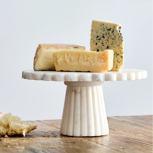 A white ceramic fluted cake stand holding several wedges of cheese, set on a rustic wooden table against a light gray wall.