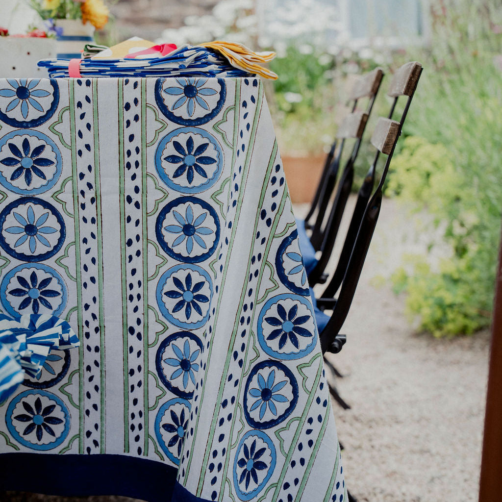 A patterned tablecloth with blue and white geometric medallion motifs draped over a table in an outdoor garden setting, with chairs and a stone wall in the background.