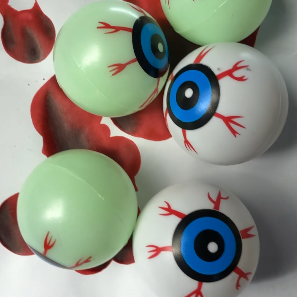 Five plastic eyeball-shaped balls, some pale green with red blood-like vessels and some white with realistic blue-and-red irises, arranged on a white surface with red splatter graphics.