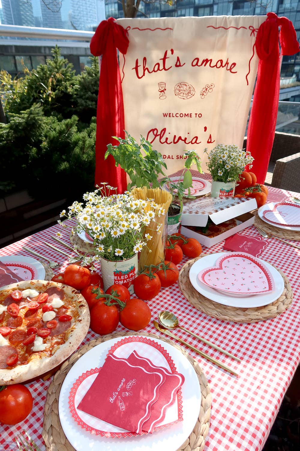 A styled outdoor table setup with a red-and-white gingham tablecloth, heart-patterned plates, red napkins, tomatoes, flowers, and a large cream banner reading 'that's amaze' and 'Welcome to Olivia's Baby Shower'.