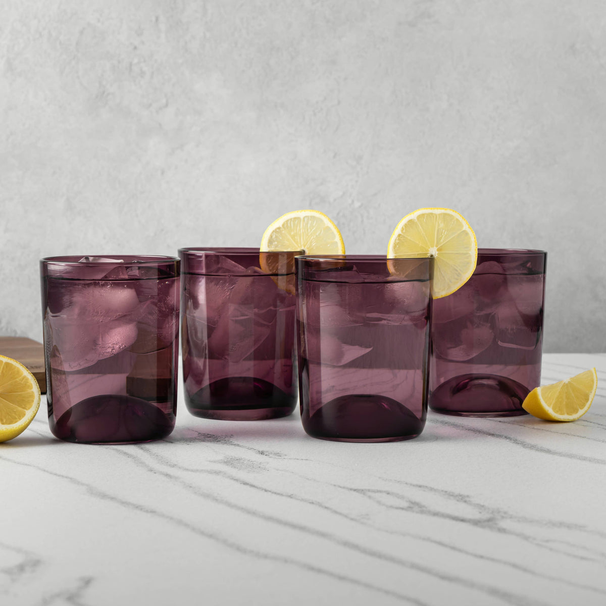 Four drinking glasses in a translucent smoky purple color, arranged on a white marble surface with lemon slices garnishing them, against a light gray textured wall.