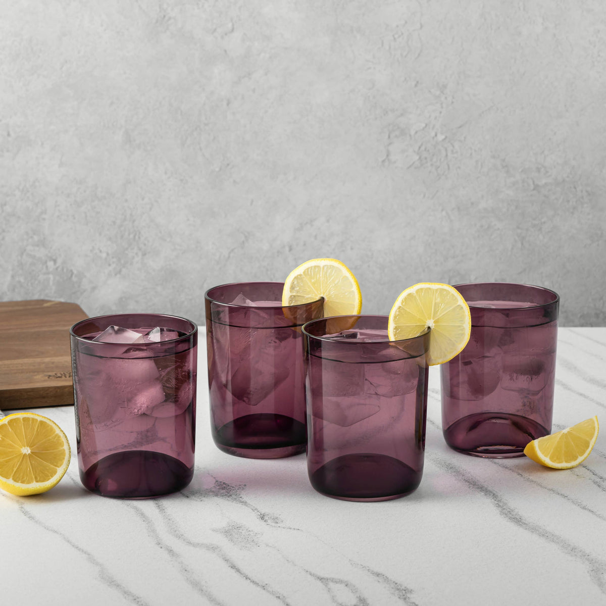 Four translucent purple drinking glasses arranged on a white marble surface with lemon slices and a folded cloth, against a light gray textured wall.
