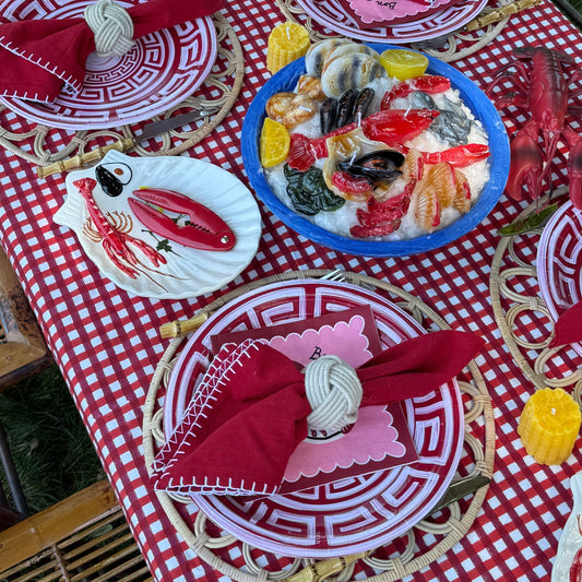 An outdoor dining table styled with a red and white checkered tablecloth, matching plates, red napkins, and a central blue platter of seafood, set on bamboo chairs with grass visible in the background.