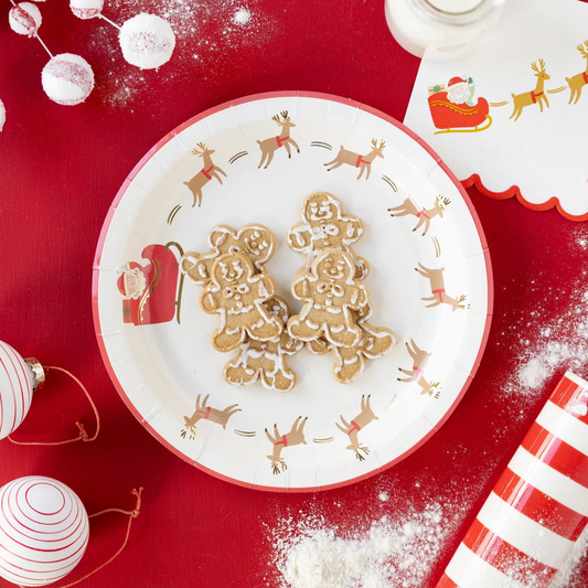 A festive holiday paper plate with a white base, red rim, and printed reindeer and sleigh motifs, styled on a red surface with gingerbread cookies and Christmas decorations.
