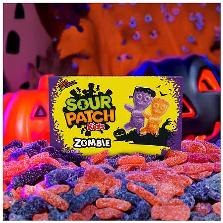 A box of Sour Patch Kids Zombie gummy candy placed in a pile of colorful sugar-coated gummies, with Halloween-themed props like a pumpkin and jack-o'-lantern in a dark, moody setting.