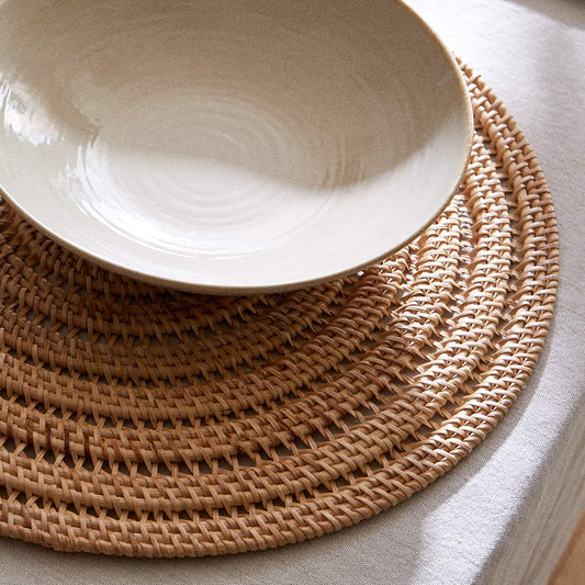 A light beige ceramic bowl with a glossy finish, resting on a round woven straw placemat over a white tablecloth.