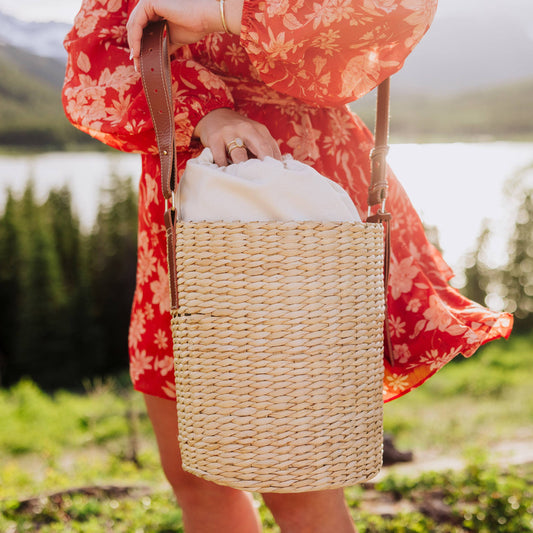 A person outdoors by a lake wearing a red floral dress and holding a large woven straw tote bag with brown leather shoulder straps.
